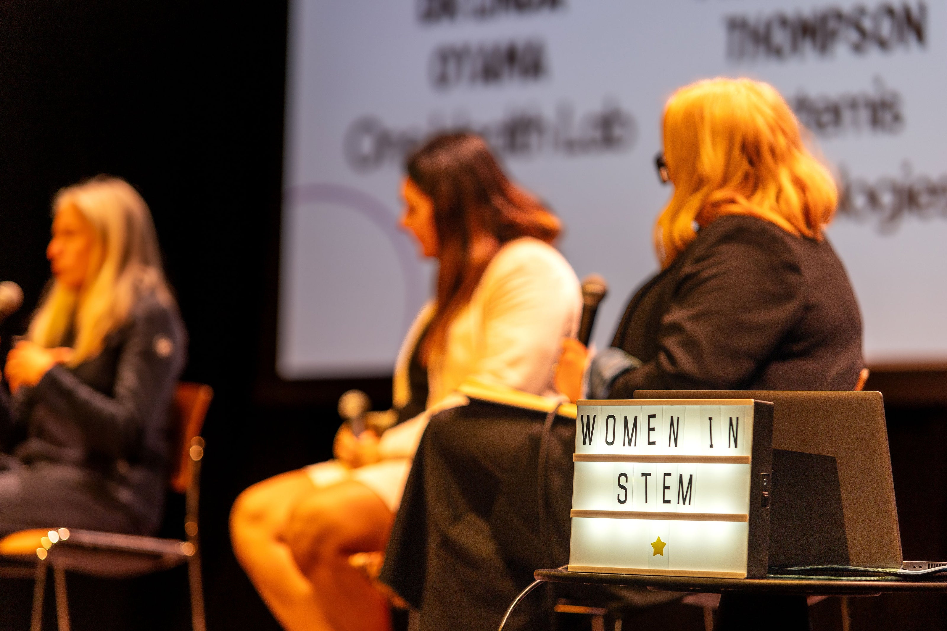 In the foreground, a lit up sign reads 'Women in STEM'. Behind it, three women sit on a stage holding microphones they all look at the woman on the left who is speaking.